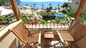 A balcony with wooden chairs and a table, overlooking a beach with umbrellas and palm trees.