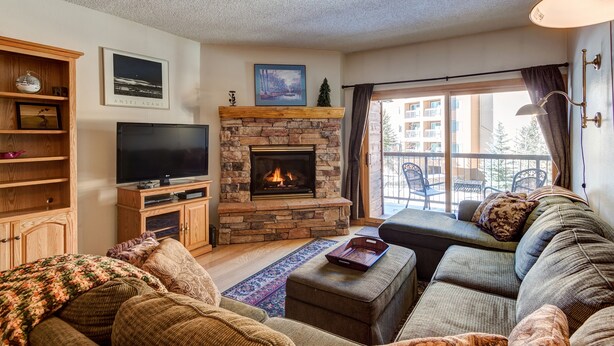 A living room with a stone fireplace, a television, a sofa, and a balcony with outdoor furniture.