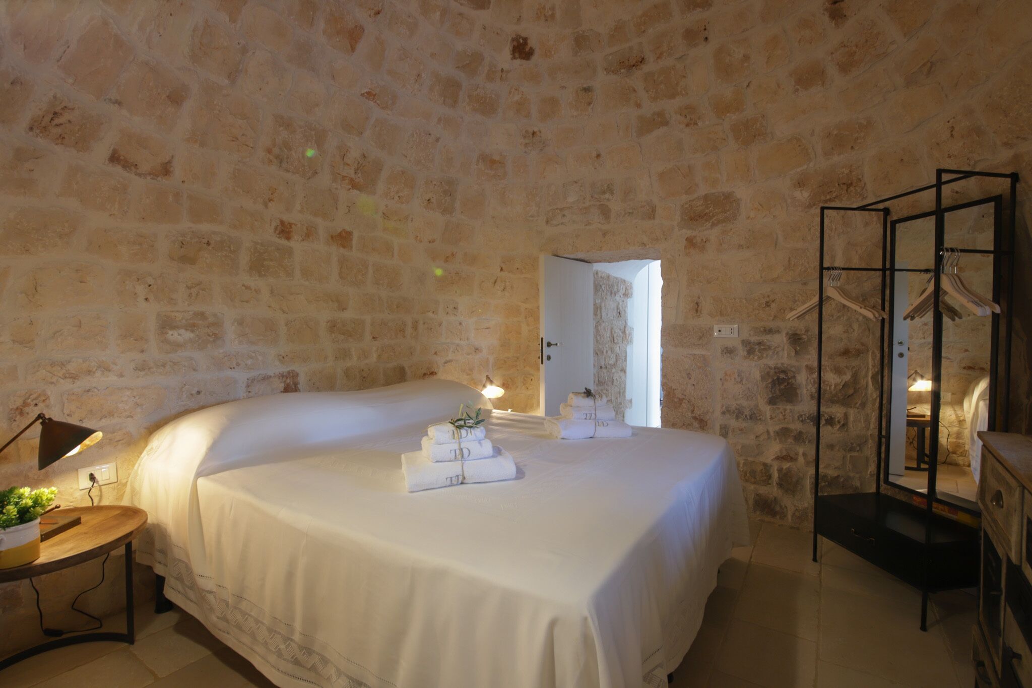 A room with a stone wall, a bed with white linens, a bedside table with a lamp, and a mirror with clothes hanging.