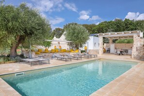 A swimming pool with lounge chairs and a pergola, surrounded by trees and a stone building.