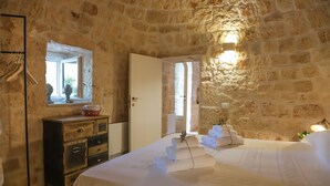 Une pièce aux murs de pierre, avec un lit, une commode, un miroir et une fenêtre.
