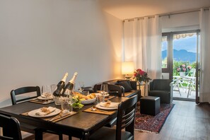 Un coin repas avec une table dressée pour deux personnes, comprenant du vin, des verres et une bouteille de champagne. On aperçoit un canapé et un fauteuil au fond, ainsi qu’une vue sur un balcon avec une table et des chaises.