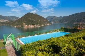 Une piscine entourée d’une clôture verte, bordée d’une végétation luxuriante et donnant sur un lac, avec des montagnes en toile de fond.