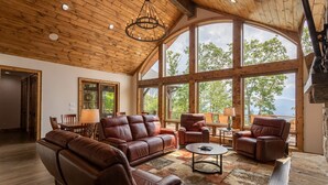A spacious living room with leather sofas, a coffee table, and large windows offering a view of trees. - Clouds Rest- luxury pet friendly home in Eagles Nest with hot tub, year round views, and more (Banner Elk)