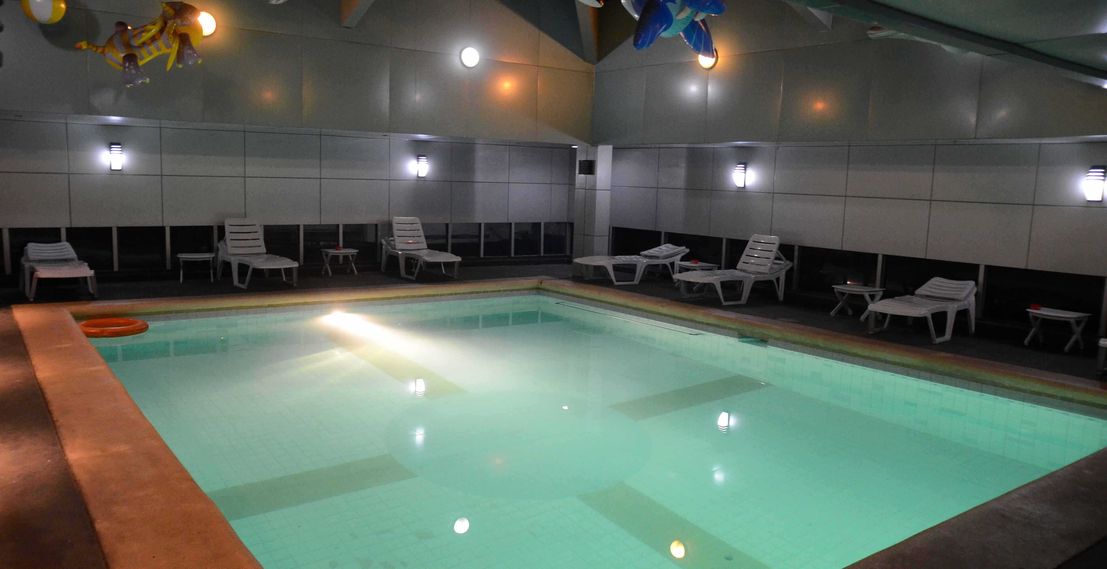 An indoor swimming pool with clear water, surrounded by white lounge chairs and small tables.