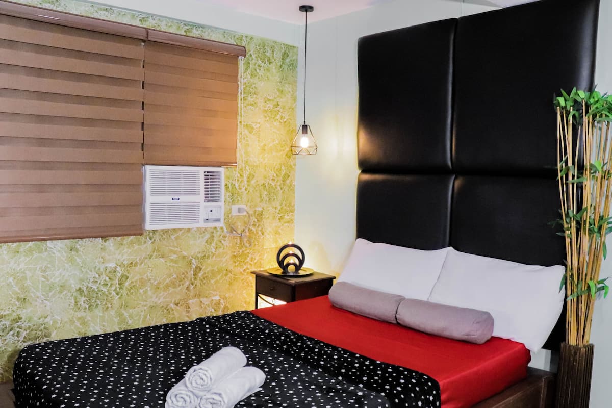 Cama bem arrumada com colcha vermelha e preta com bolinhas, um travesseiro branco e uma toalha branca enrolada. Há uma cabeceira preta, uma pequena mesa de cabeceira com uma luminária e um ar condicionado fixado na parede.