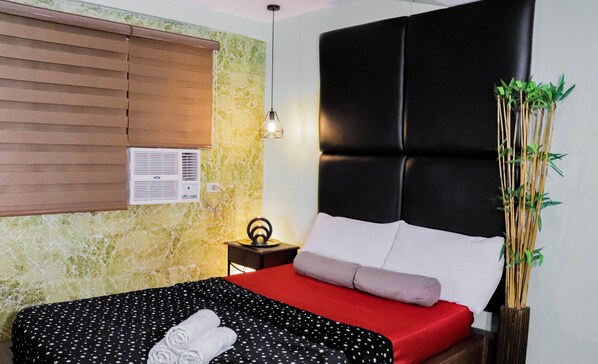 A neatly made bed with a red and black polka dot bedspread, a white pillow, and a rolled white towel. There is a black headboard, a small bedside table with a lamp, and a wall-mounted air conditioner. - Sprout Rooms (Quezon City)