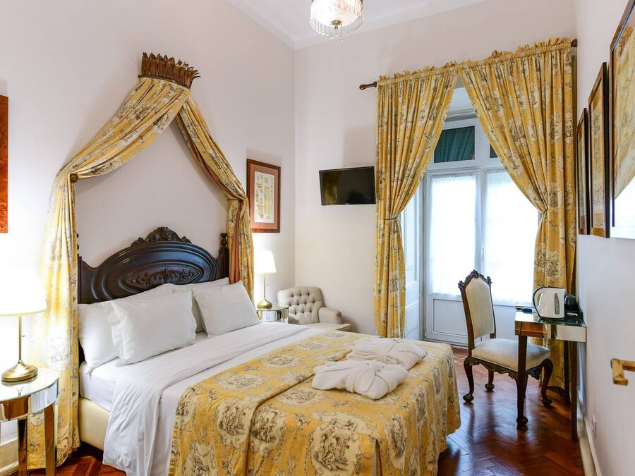 A four-poster bed with a canopy, a bedside table with a lamp, a chair, a desk with a computer, and a window with curtains.