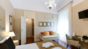 A hotel room with a bed, a sitting area with a sofa and armchairs, a desk with a chair, a TV mounted on the wall, and a chandelier. - Casa de Sao Mamede Hotel (Lisbon)