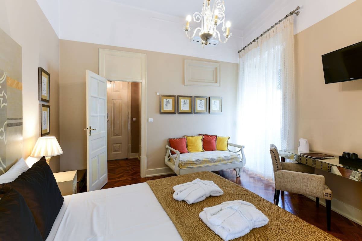 A hotel room with a bed, a sitting area with a sofa and armchairs, a desk with a chair, a TV mounted on the wall, and a chandelier.