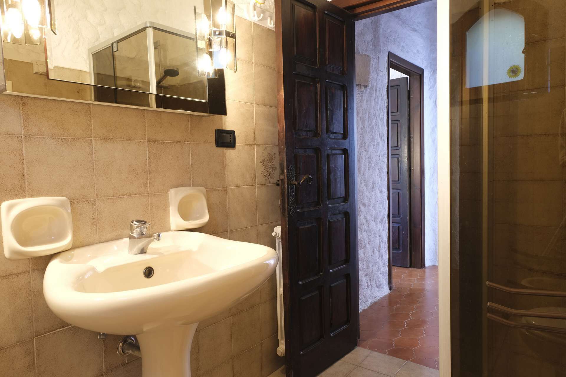 A bathroom with a white sink, two urinals, a dark wooden door, and a mirror with lights.