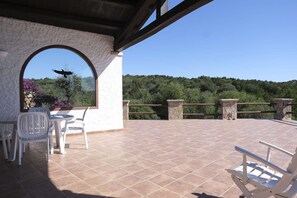 A covered patio with a stone floor, white chairs, and a view of a forested hillside. - Aria di Vacanza - Costa Dorata Cod 4 (Loiri Porto San Paolo)