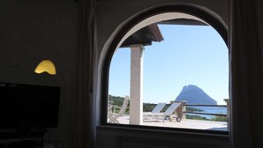 Een kamer met een groot, rondboogvenster met uitzicht op een zwembadgedeelte met ligbedden en een berg op de achtergrond.