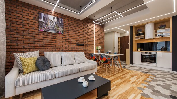 A modern living room with a brick wall, a sofa, a coffee table, and a kitchen area with wooden cabinets and a dining table. - P&O Apartments Cybernetyki 6 (Warsaw)