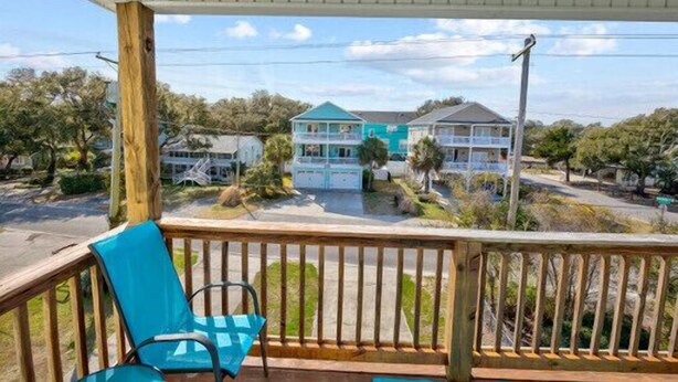 A covered porch with a wooden railing, a blue chair, and a view of a turquoise house with a white roof.