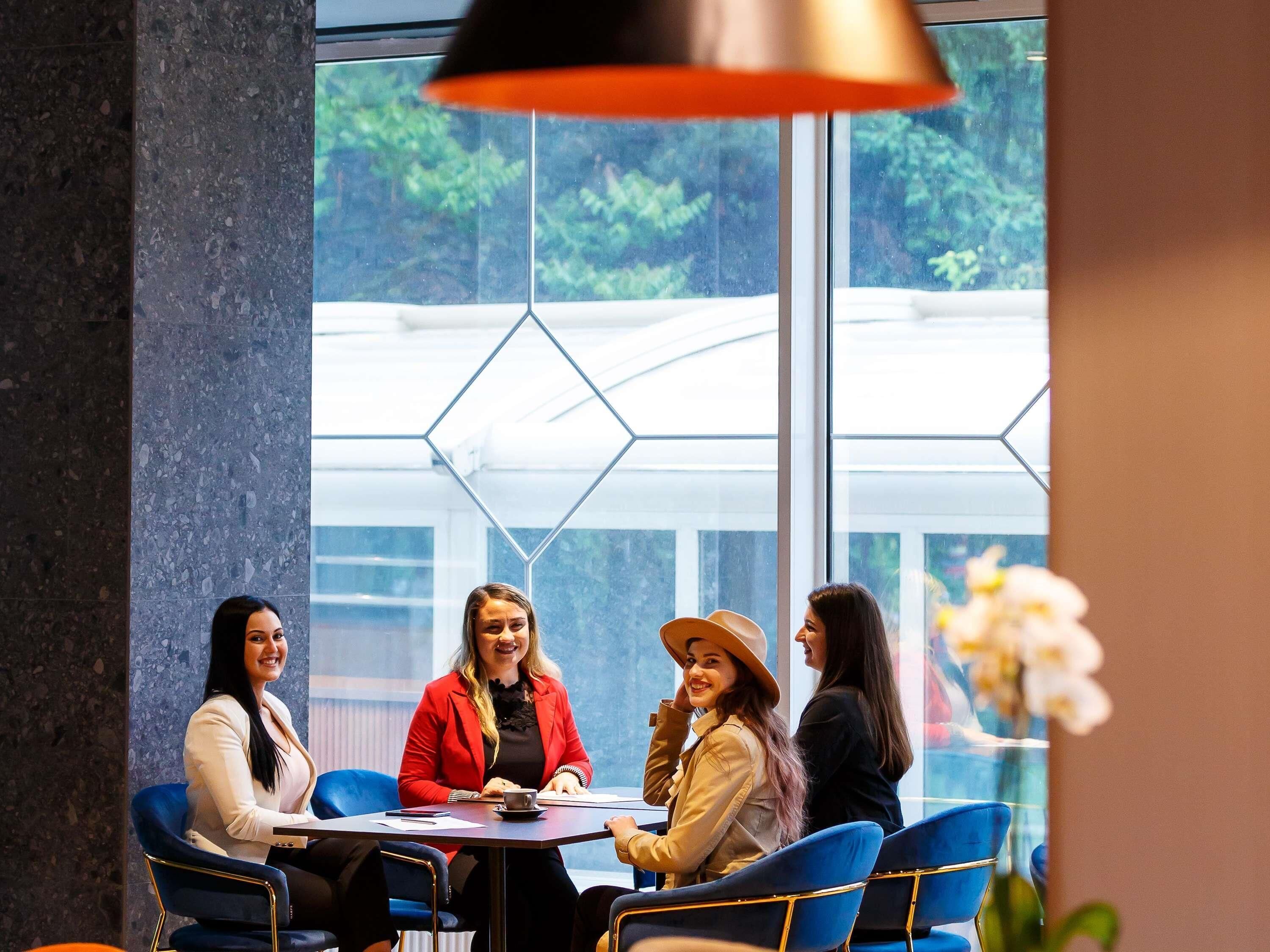 A group of people seated around a table in a modern indoor setting with large windows and geometric glass panels.