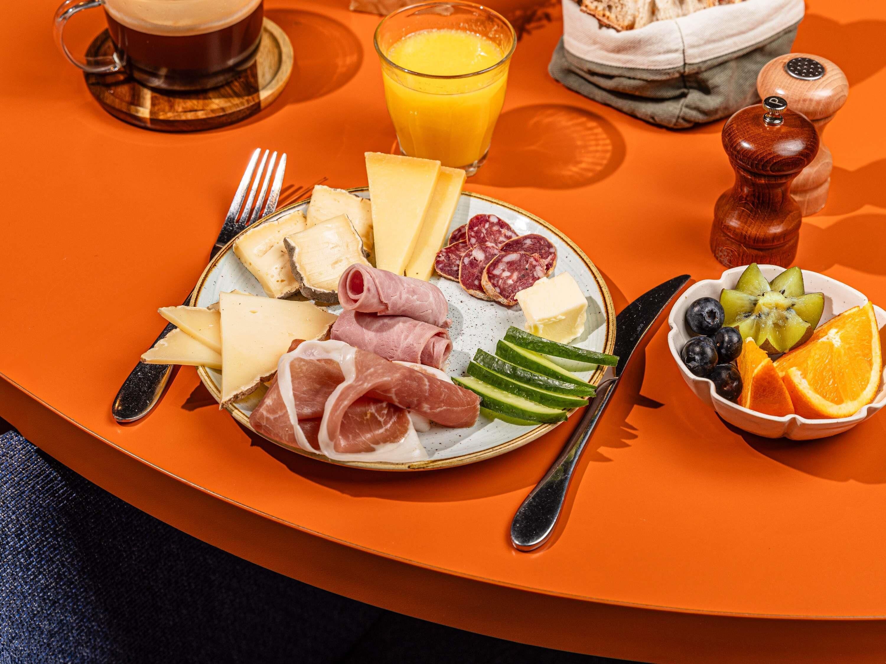 A breakfast setting with a plate of cheese, meats, and fruits, a glass of orange juice, and a cup of coffee on an orange table.