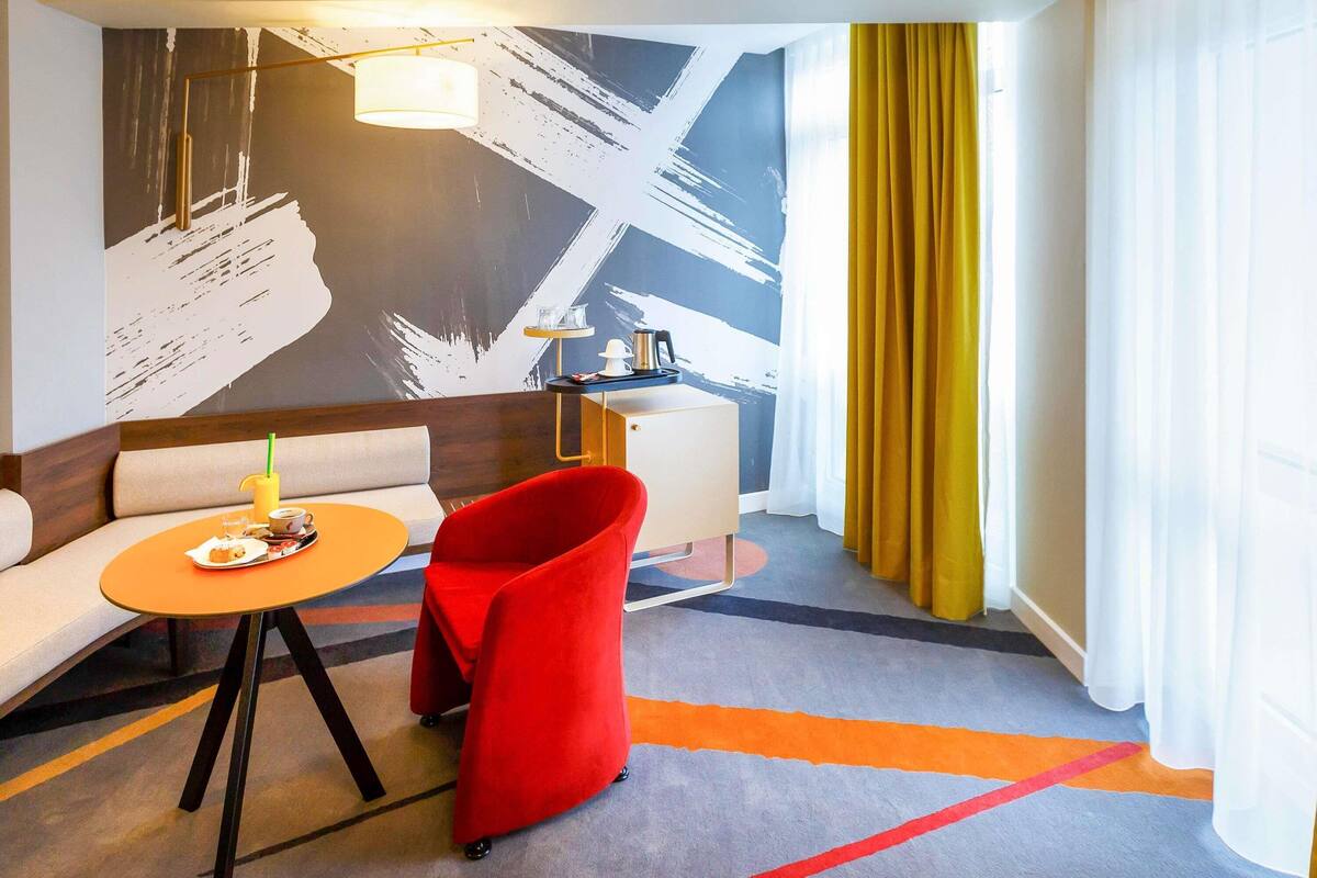 Una habitación de hotel moderna con un sofá, una silla roja, una pequeña mesa con una bebida, un escritorio y un cuadro abstracto grande en la pared.