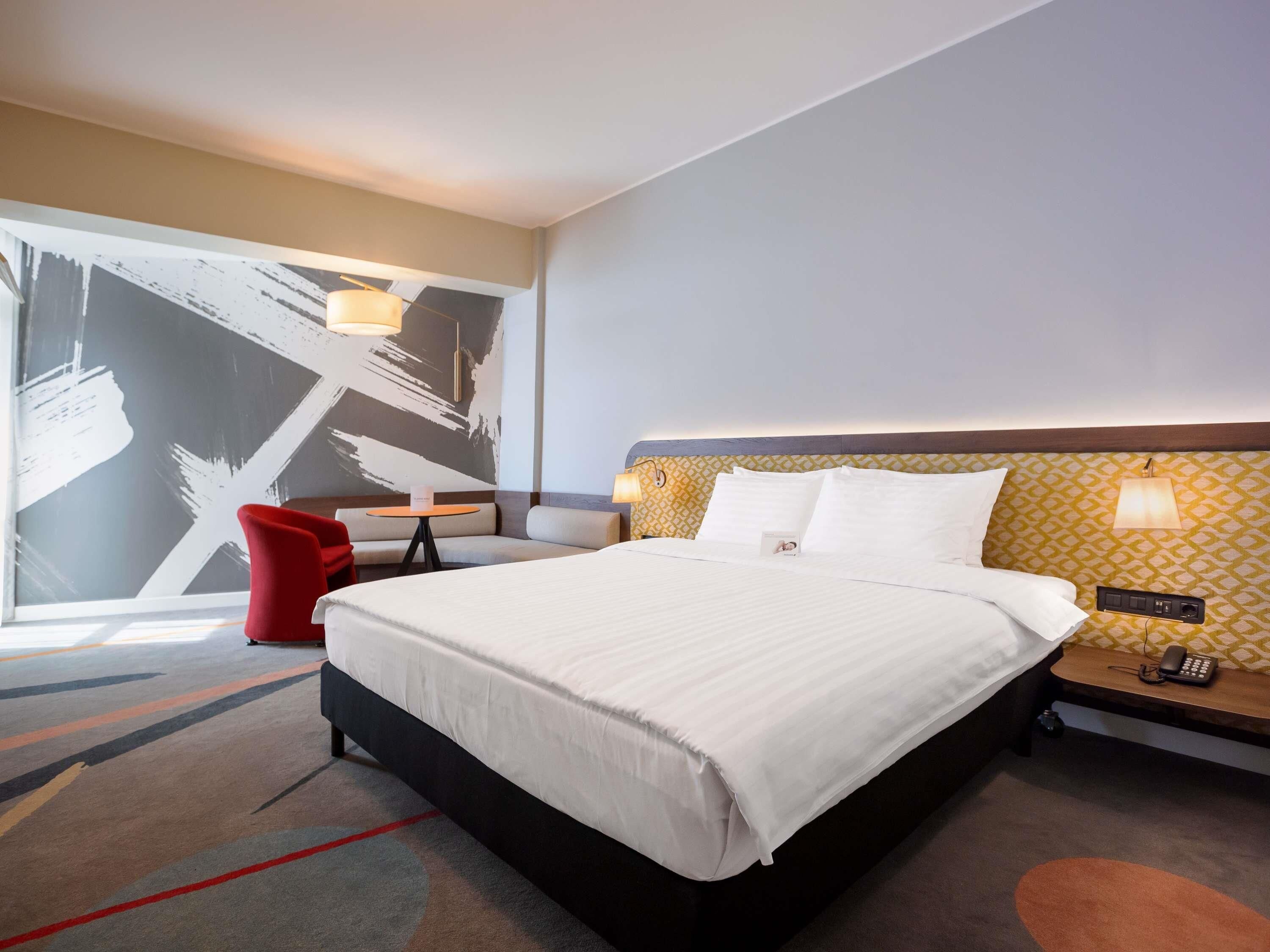 A hotel room with a large bed, a red chair, a small table, and a wall with a geometric pattern.