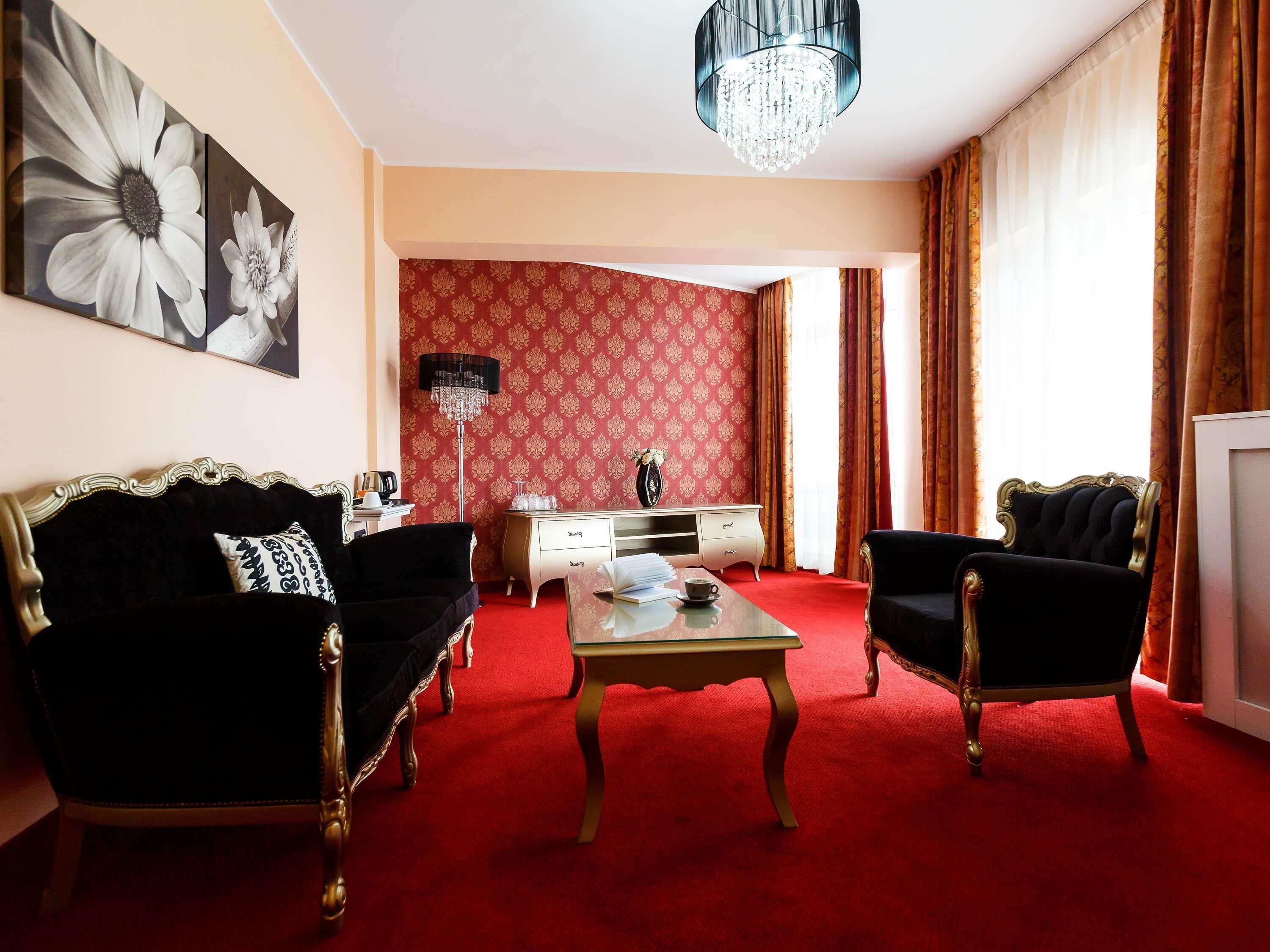 Una habitación con alfombra roja, dos sofás negros, un aparador blanco, una araña y un cuadro grande con flores en la pared.
