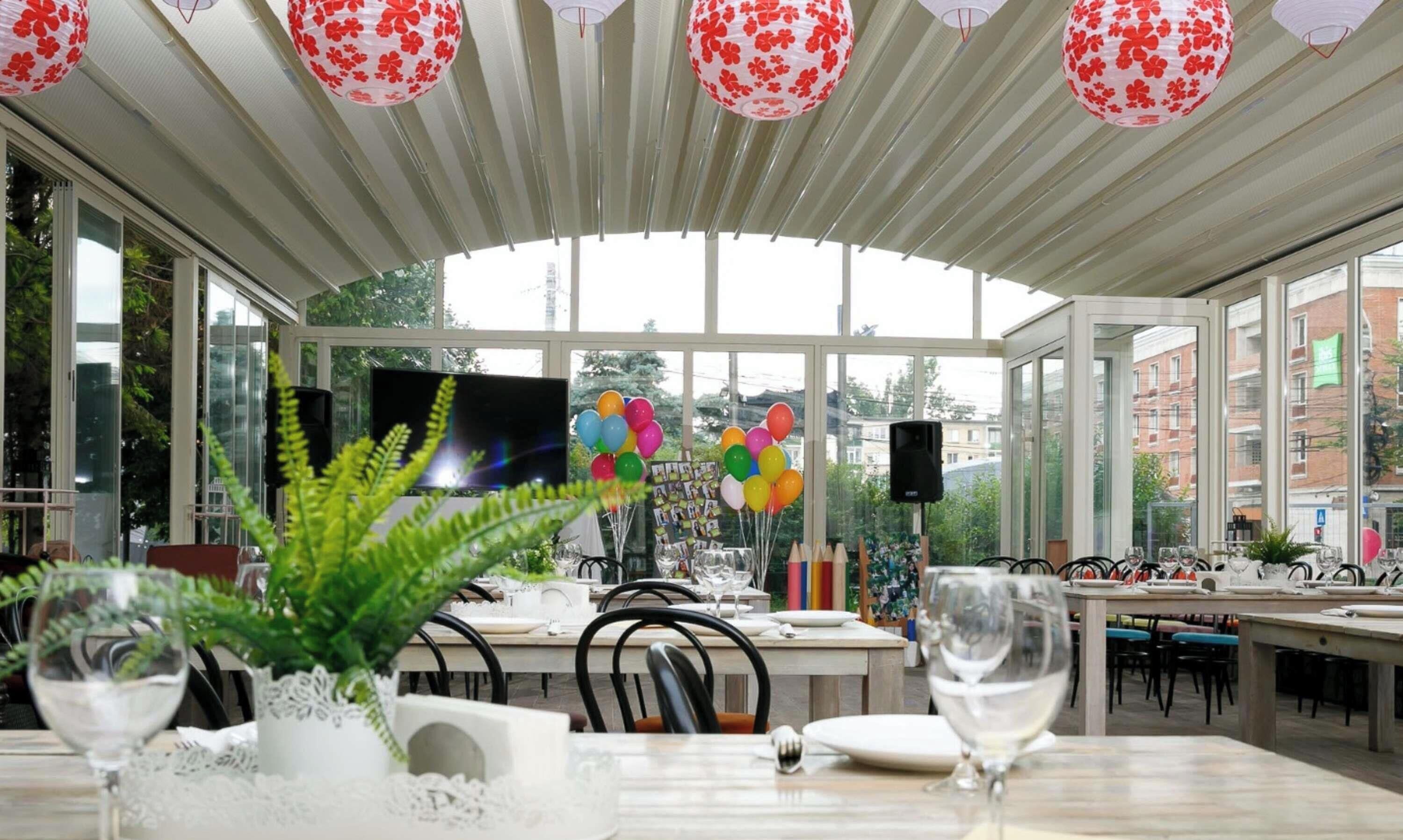 A restaurant with a glass enclosure, tables set for dining, and red floral lanterns on the ceiling.