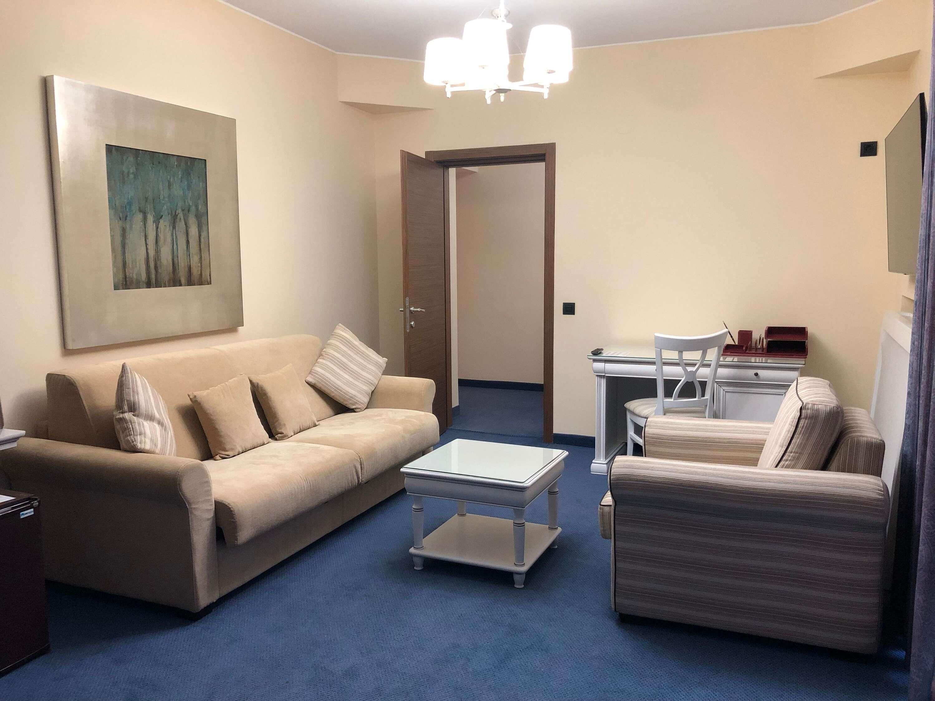 A living room with a sofa, a coffee table, and a chair. There is a painting on the wall and a door leading to another room.
