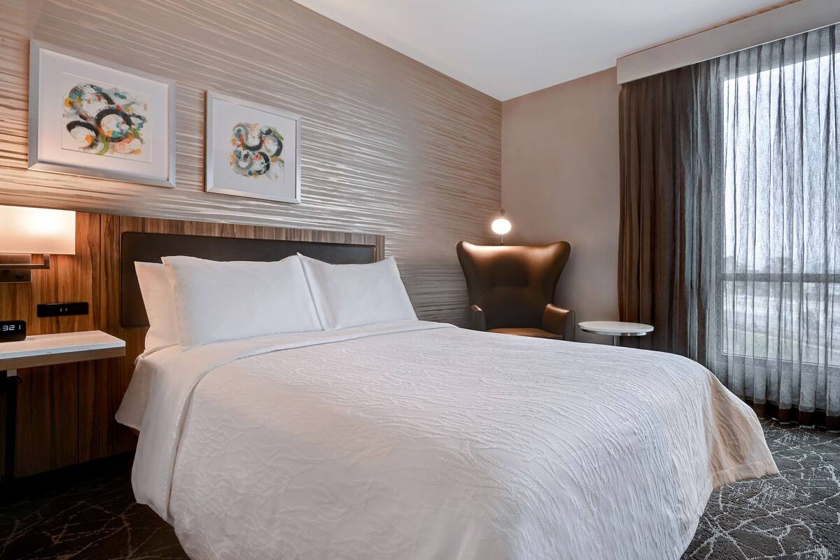 A hotel room with a large bed, a wooden headboard, a side table, a chair, and two framed artworks on the wall.