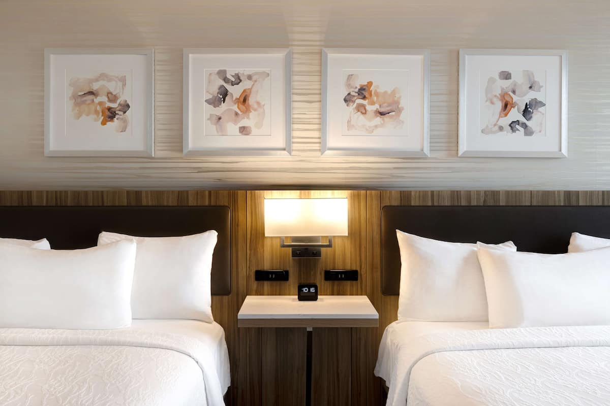 A hotel room with two beds, a nightstand, a wall-mounted lamp, and three abstract paintings above the beds.