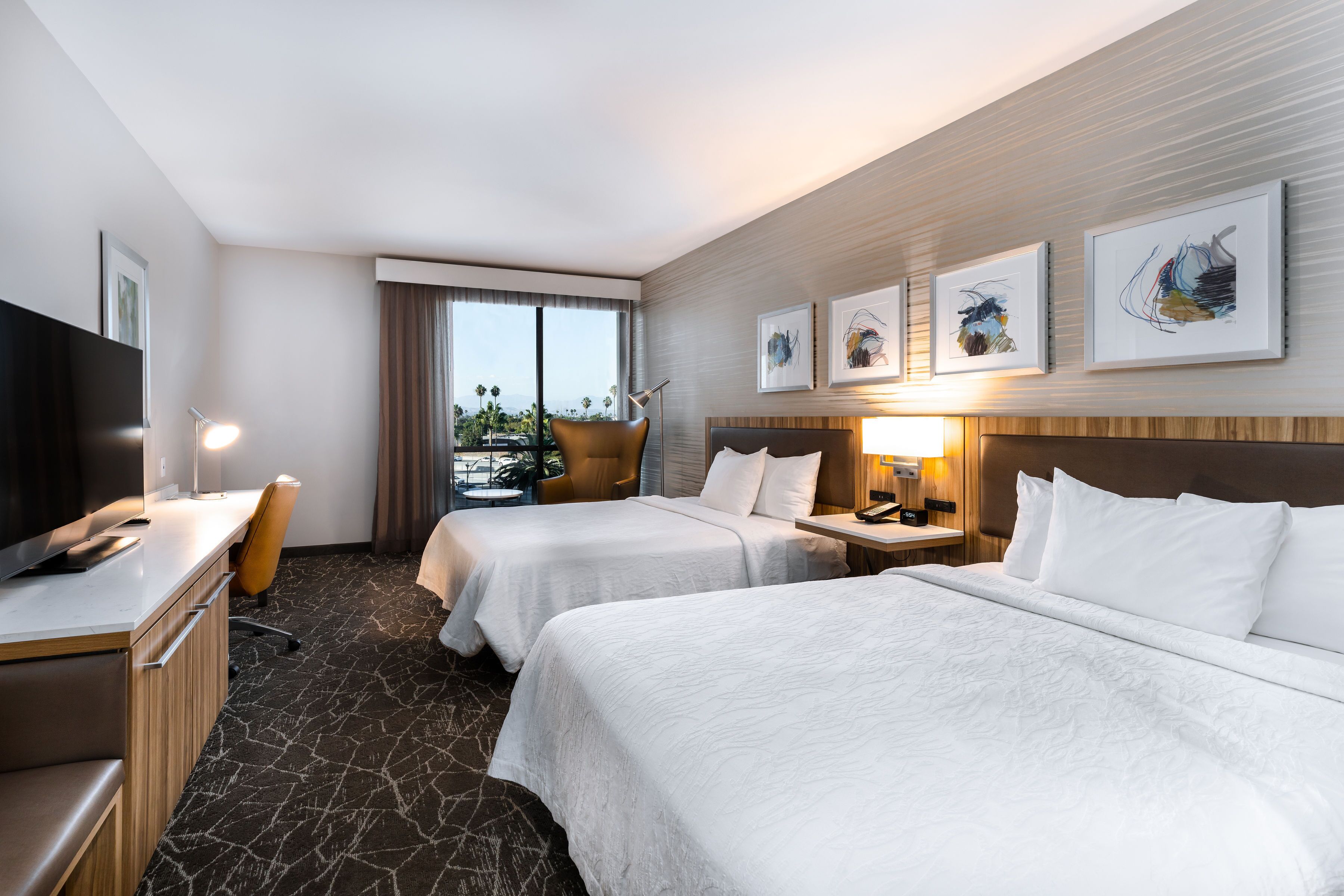 Quarto de hotel com duas camas, uma escrivaninha, uma cadeira, televisão e obras de arte na parede.