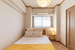 A neatly arranged bedroom with a bed, a bedside table with a lamp, a window with curtains, and an air conditioning unit.