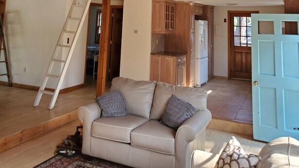 Un espacio tipo loft con techo de madera, un sofá blanco, una escalera y una zona de cocina con armarios de madera.