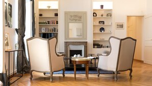 A room with two armchairs facing a fireplace, a small round table, and a bookshelf with various items. - Monceau Chic I Charming 1 Bed Apt in the 8th (Paris)