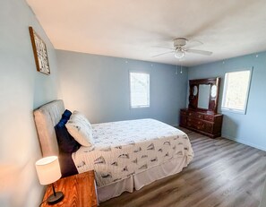 Ein Schlafzimmer mit Bett, Kommode, Deckenventilator und zwei Fenstern.