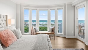 Un dormitorio con un ventanal grande con vistas a la playa, una cama con almohadas mullidas y un ventilador de techo.
