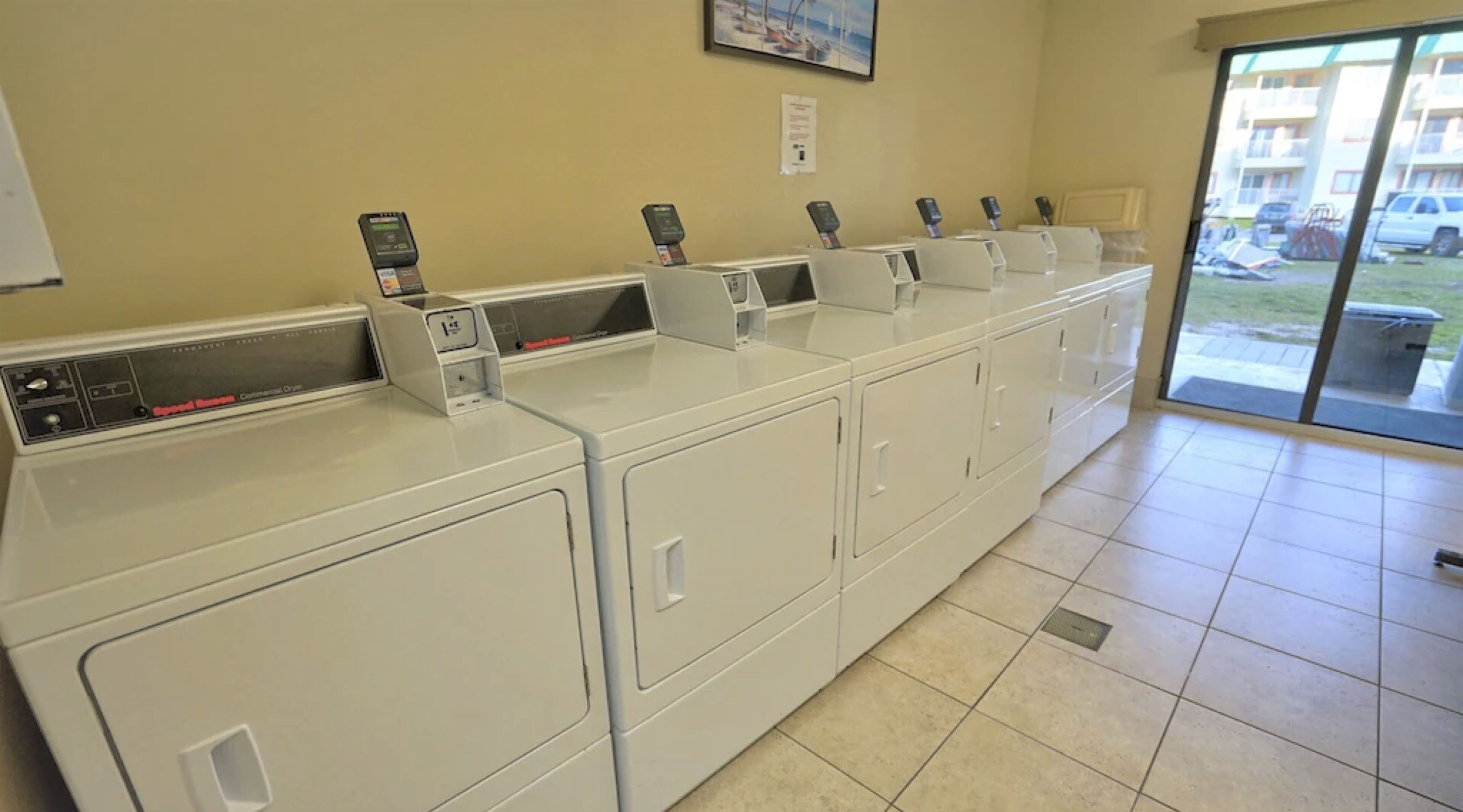 A row of white commercial washers with control panels, a tiled floor, and a view of parked cars outside through a glass door.