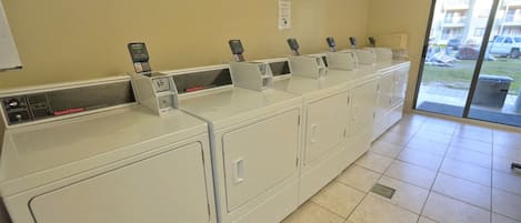 A row of white commercial washers with control panels, a tiled floor, and a view of parked cars outside through a glass door.