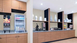 A modern kitchen area with wooden cabinets, a refrigerator stocked with beverages, and a counter with various kitchen appliances and a decorative vase.