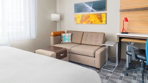 A hotel room with a bed, a sofa, a desk with a chair, and two artworks on the wall.