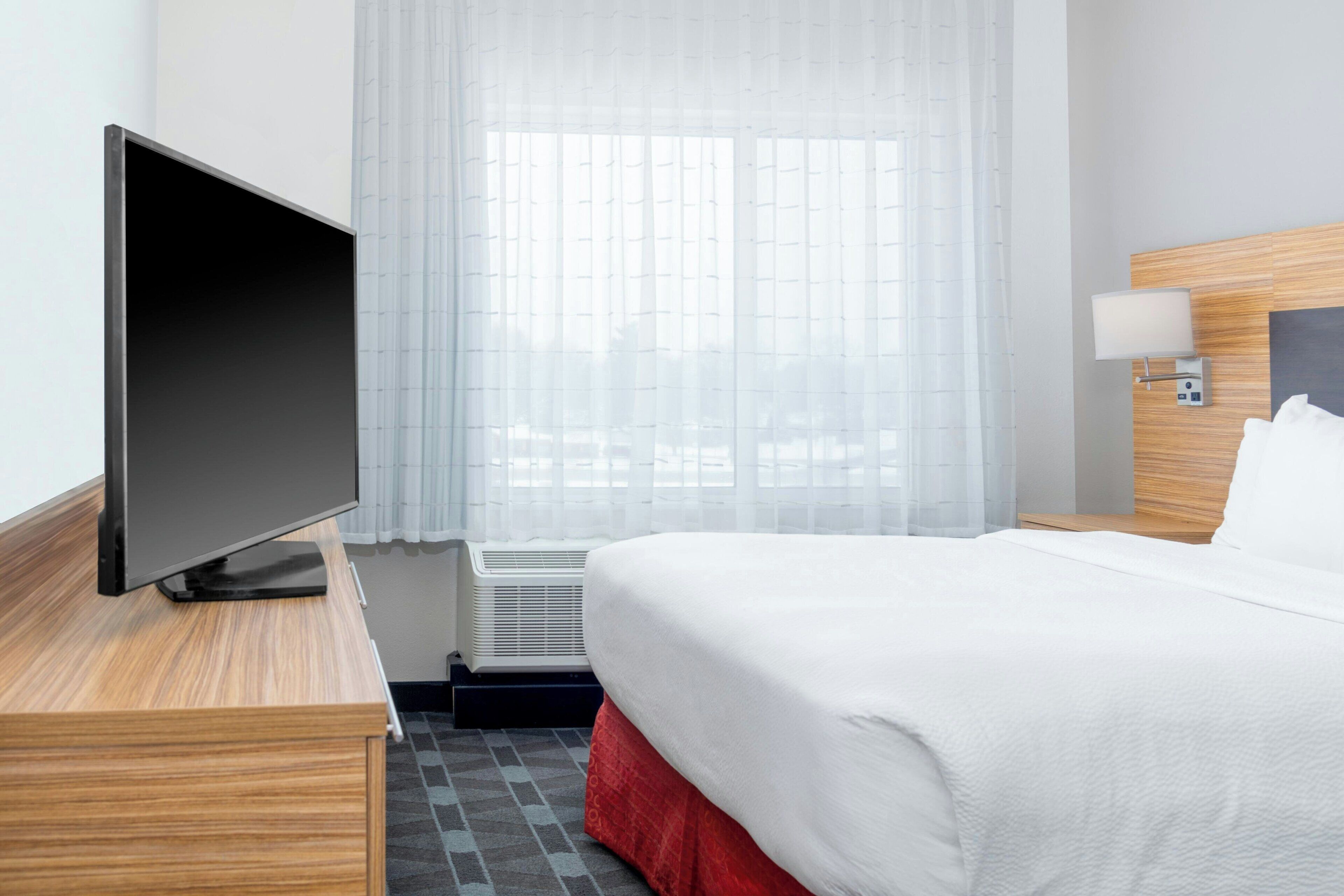 Une chambre d’hôtel avec un lit, un téléviseur posé sur une table de chevet en bois, une fenêtre avec des rideaux légers et une lampe fixée au mur.