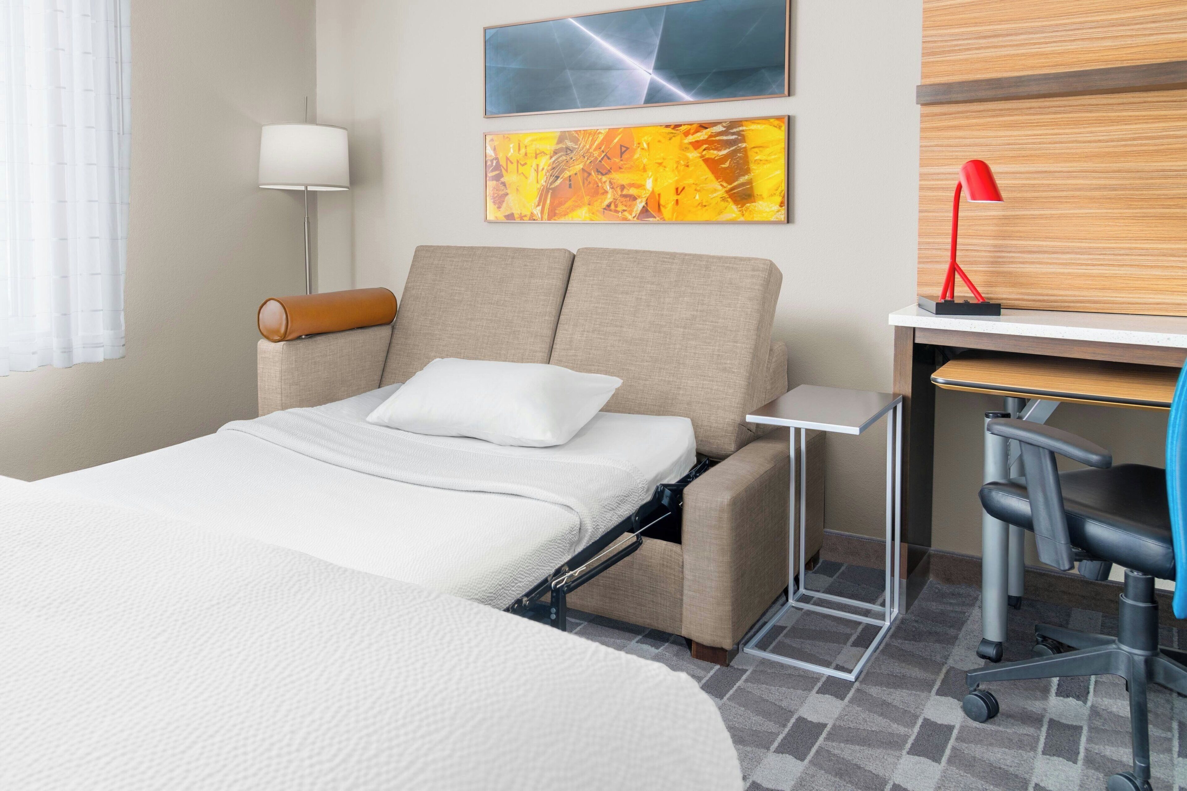 A hotel room with a bed, a desk, a chair, and two paintings on the wall.