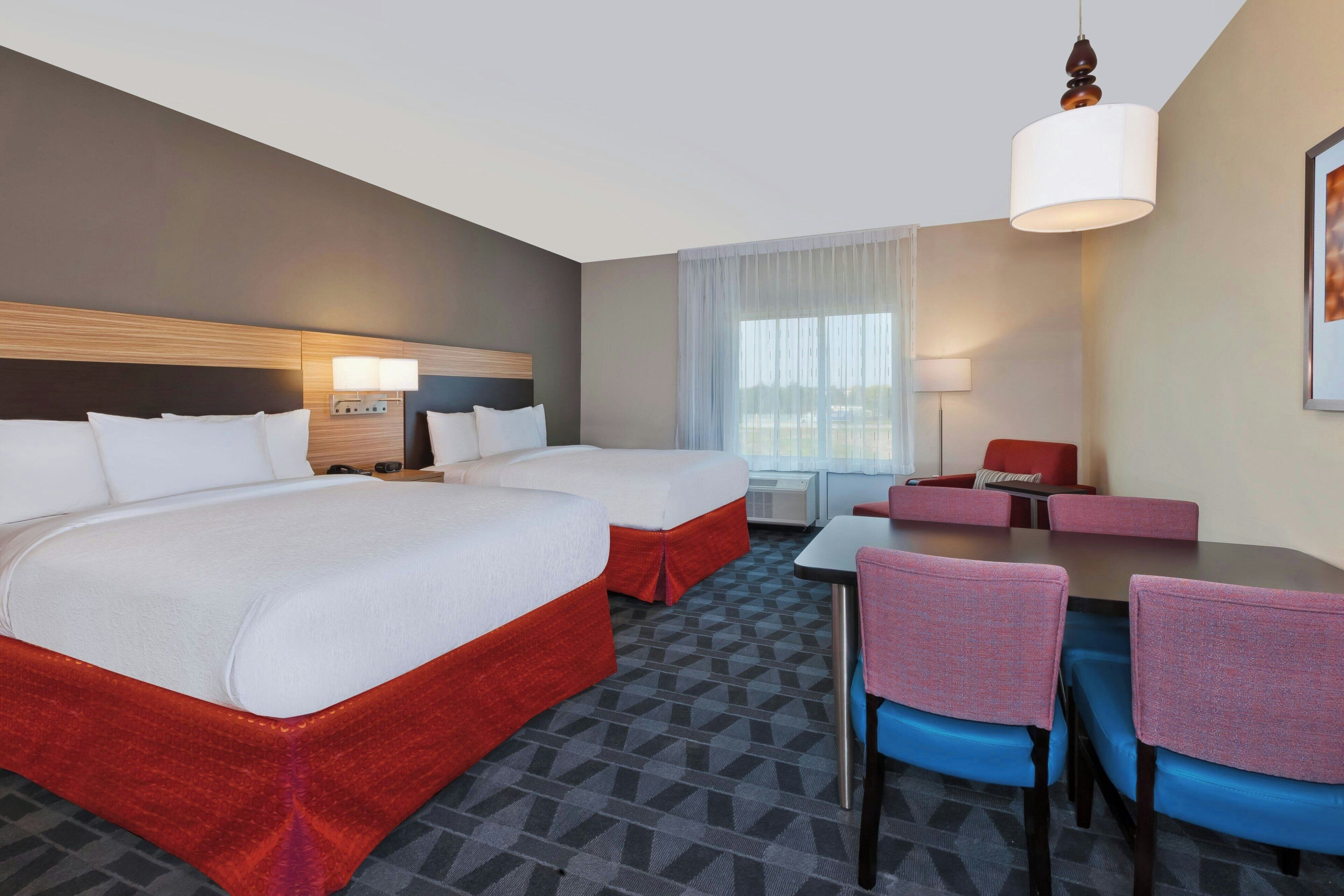 A hotel room with two beds, a desk, chairs, a lamp, and a window with curtains.