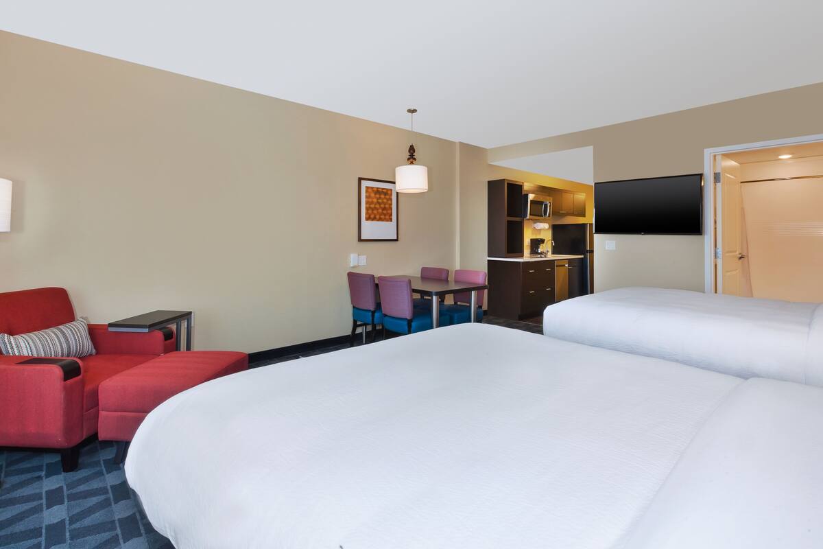 Una habitación de hotel con una cama grande, un sillón rojo, una mesita, un televisor y una kitchenette al fondo.