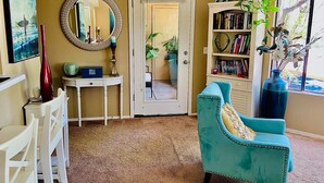Una sala de estar con un sillón azul turquesa, una mesita auxiliar blanca, una estantería y un espejo.