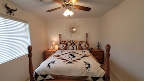 Une chambre à coucher avec un lit en bois, un ventilateur de plafond, un miroir fixé au mur et une fenêtre avec des stores.