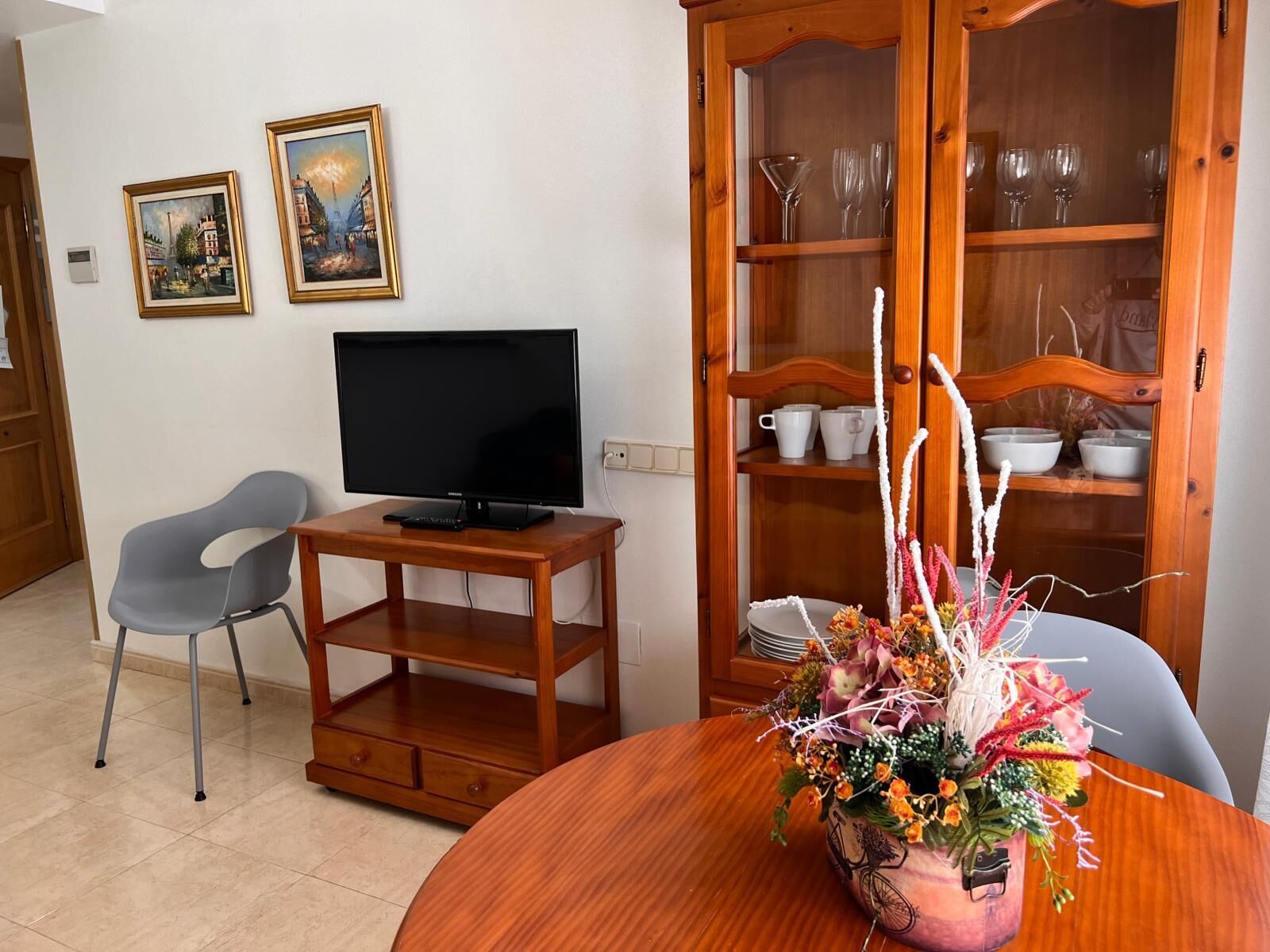 A living room with a flat-screen TV on a wooden stand, a grey chair, a dining table with a vase of flowers, and a wooden cabinet with glass doors containing white cups and bowls.