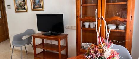 A living room with a flat-screen TV on a wooden stand, a grey chair, a dining table with a vase of flowers, and a wooden cabinet with glass doors containing white cups and bowls.