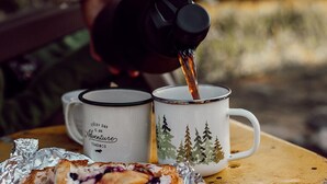 Una persona sirve una bebida caliente en una taza con diseño de bosque, junto a otra taza y un alimento sobre una mesa.