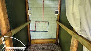 Un espacio reducido y cerrado con vigas de madera y pared de azulejos, que incluye una zona de ducha con un tubo rojo y uno azul, y un cortinero blanco.