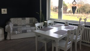 A room with a white sofa, a dining table with chairs, a window with a view of the outdoors, and a radiator under the window. - Vacation apartment directly on the Ems dyke in an idyllic location (Westoverledingen)