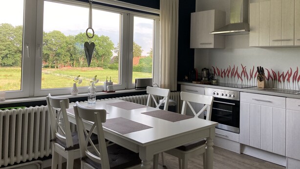 A modern kitchen with white cabinetry, a dining table with chairs, and a view of a garden through large windows. - Vacation apartment directly on the Ems dyke in an idyllic location (Westoverledingen)